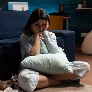 imagem com uma mulher, morena, sentada no chão da sala, encostada no sofá, de pijama, com o travesseiro no colo, com aparente depressão